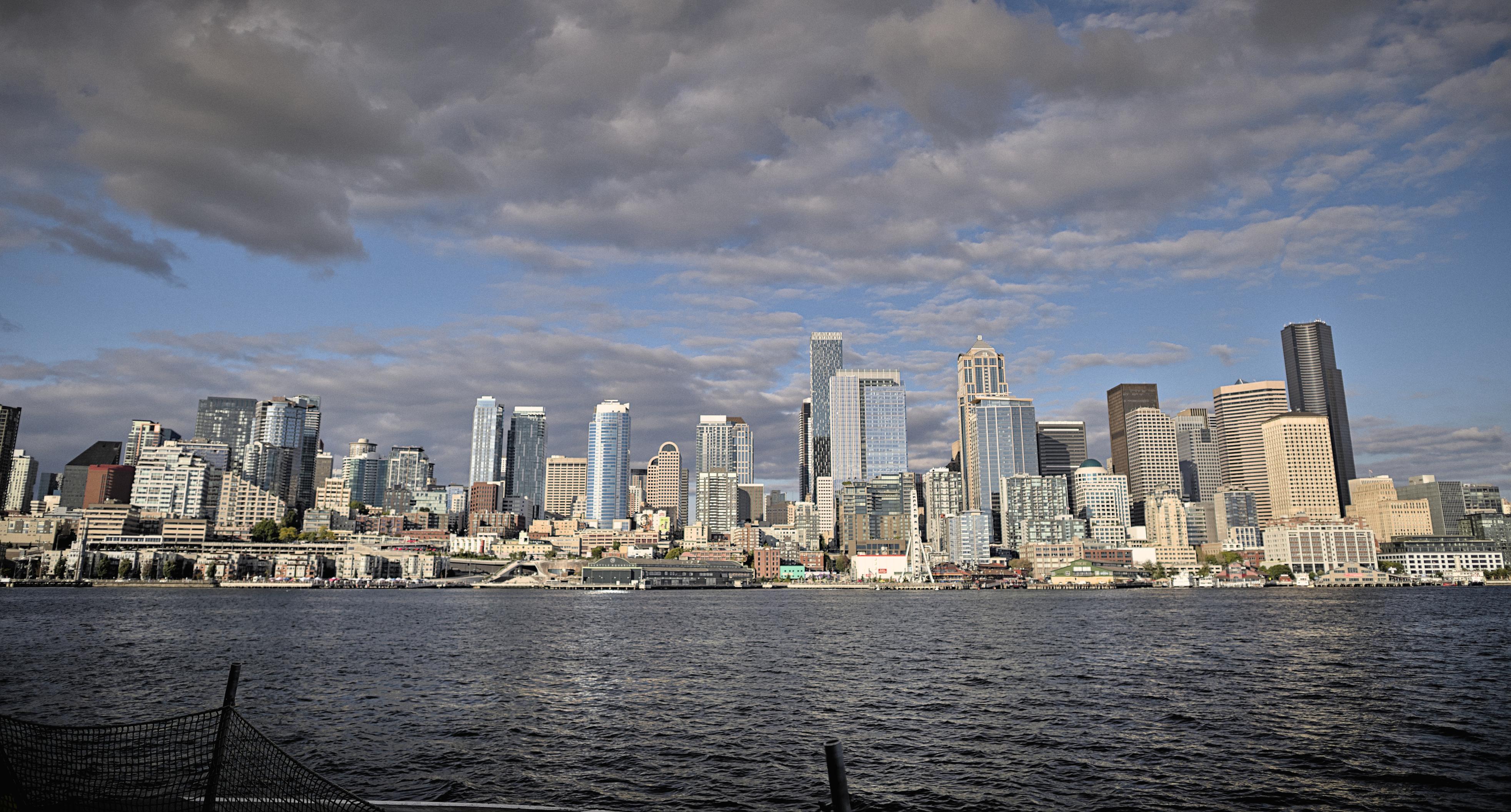 very wide shot of the Seattle skyline taken from the deck of a ferry in Elliott Bay. The water is mostly calm and the cloudy skies above show about %50 coverage.