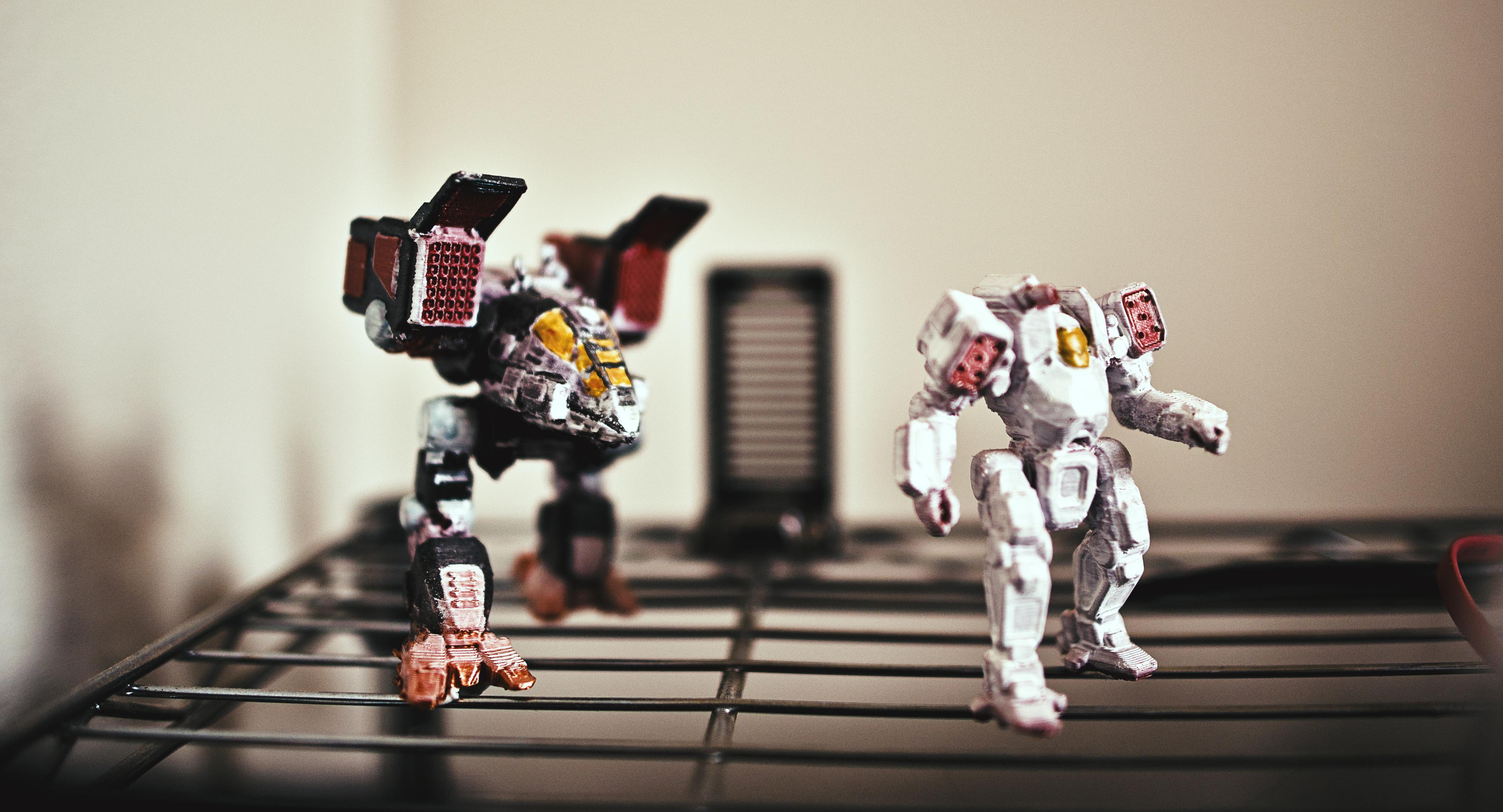 macro shot of two 3D-printed, partially painted battle tech figures. On the left is a CPT-C1 with black, red, and white marks and on the right is an all white ACH-Prime with some red on the shoulder mounts. The background is flat white and the ground is a shiny metal grid