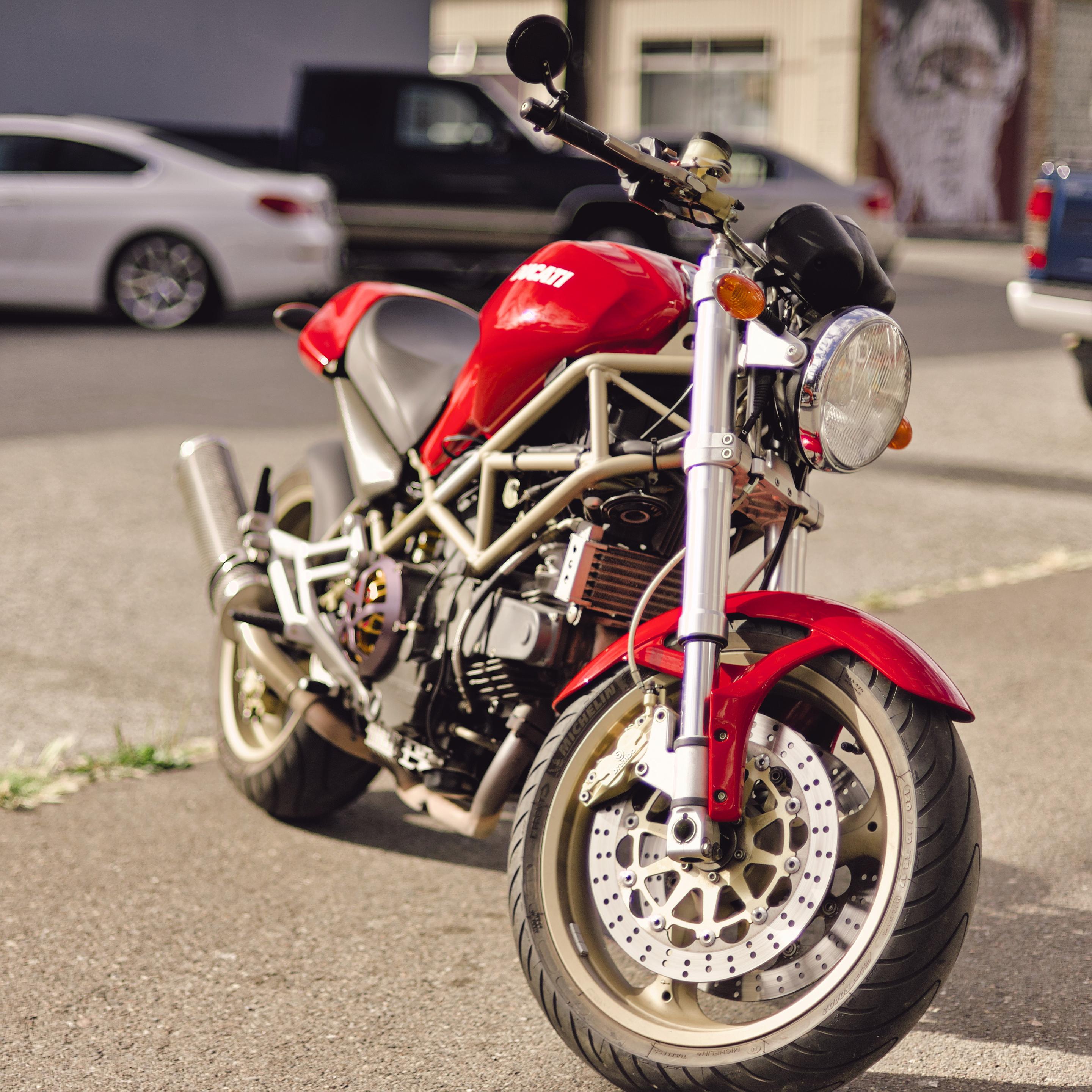 3/4 profile shot of a very Red Ducati M900 model year 2000 parked on the side of the road with a few cars and a blurry mural in the background